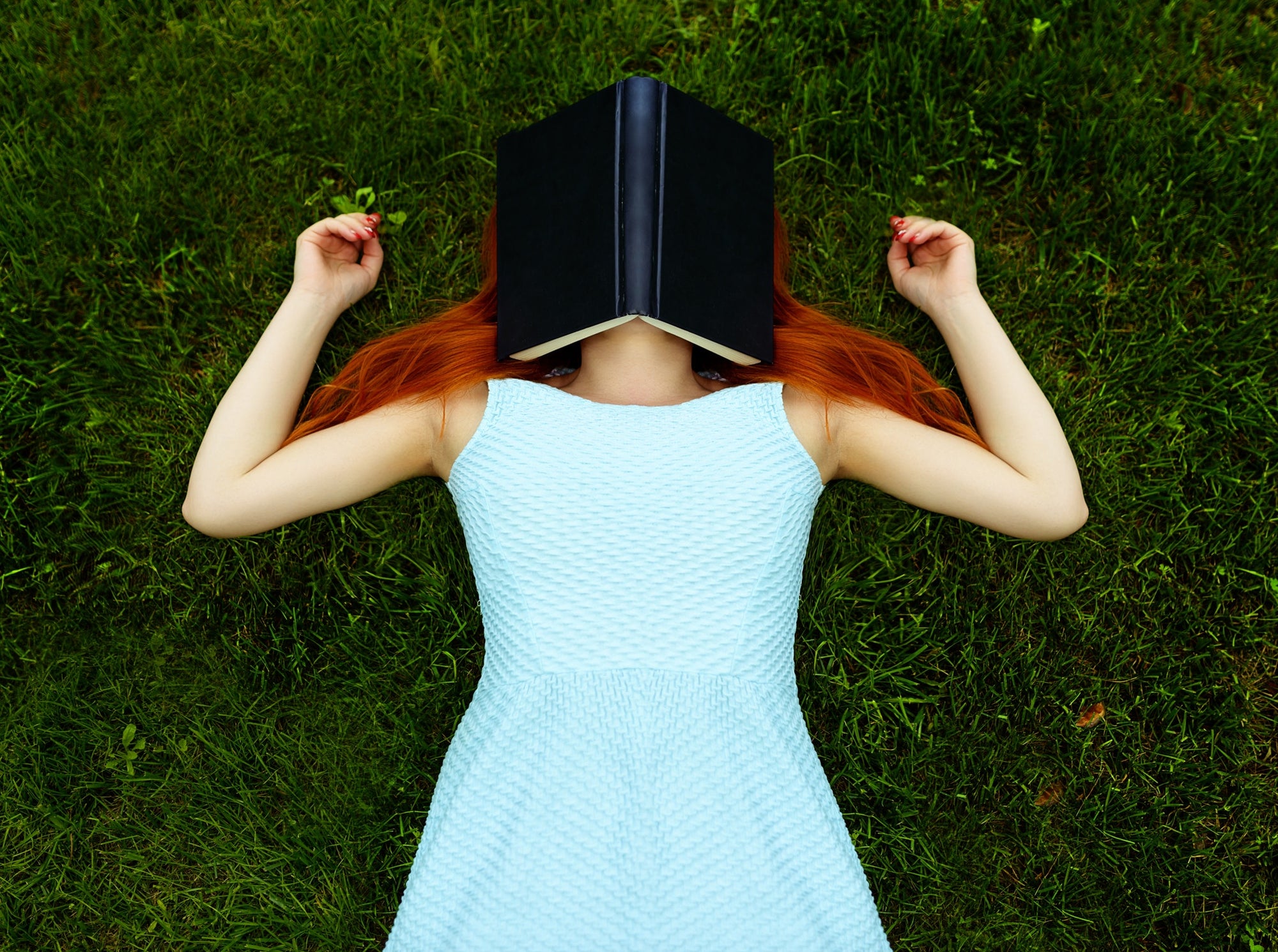 young red head woman lying on field with book covering her face.