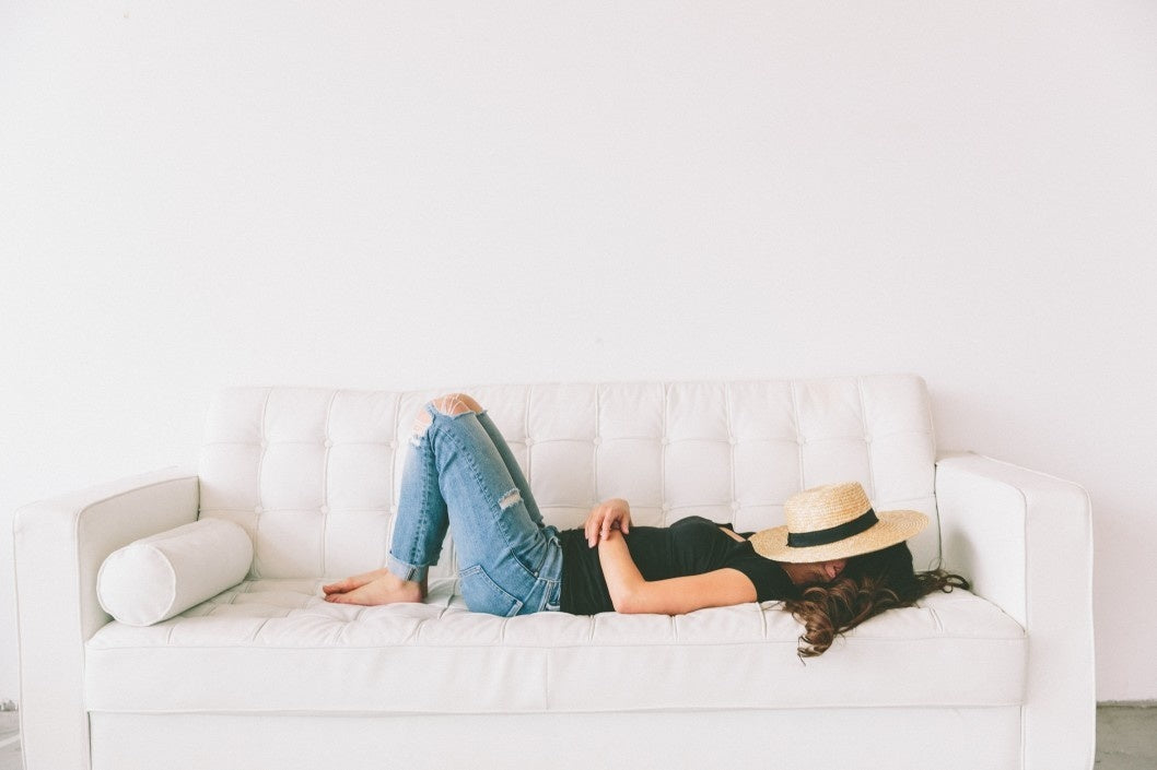 young woman sleeping on a bright white couch with a hat covering her face.