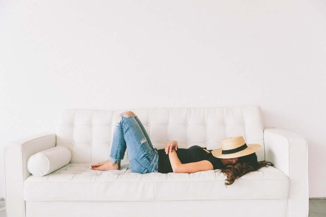 young woman sleeping on a bright white couch with a hat covering her face.