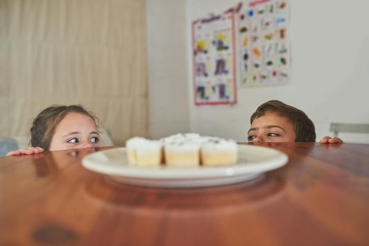 little girl and boy hiding behind table at kitchen and looking at cupcakes kept on table