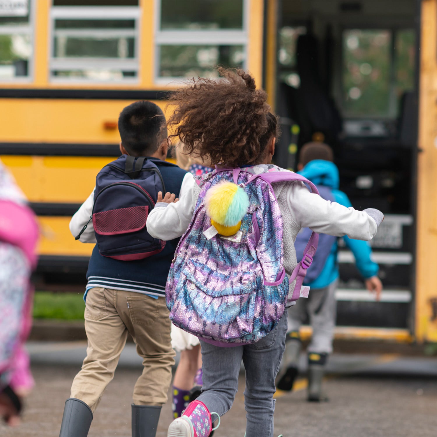 A child with curly hair walks away from the camera wearing a colorful tie-dye backpack with a fluffy yellow and blue pom-pom, alongside other children carrying backpacks near a yellow school bus.