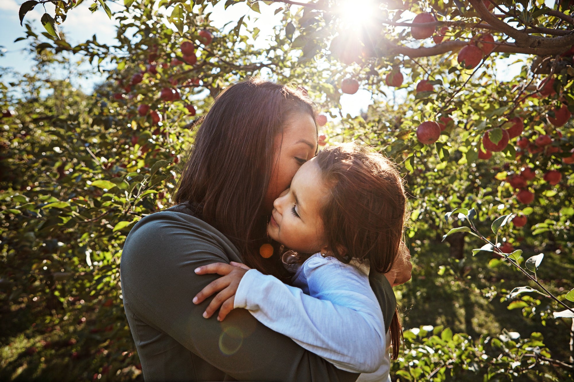 mother is kissing her daughter on the cheek, apple trees on the background