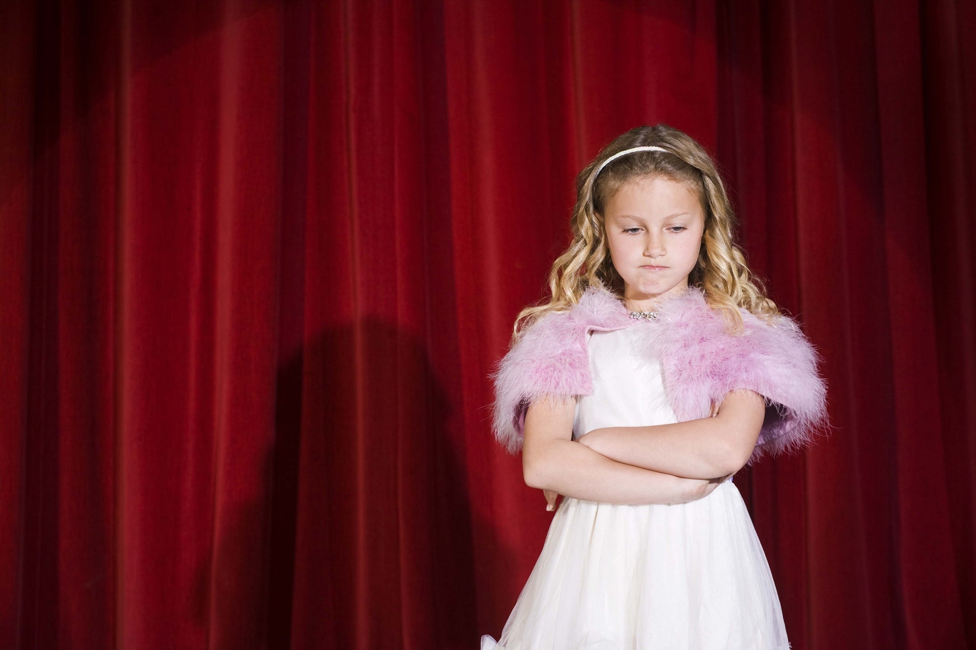 Front view of young girl standing in spotlight against red stage curtain looking annoyed