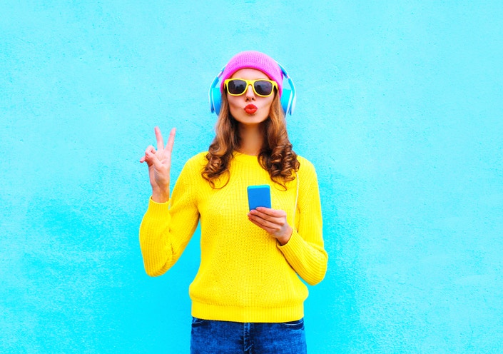 beautiful girl in headphones holding mobile showing peace sign isolated on blue background