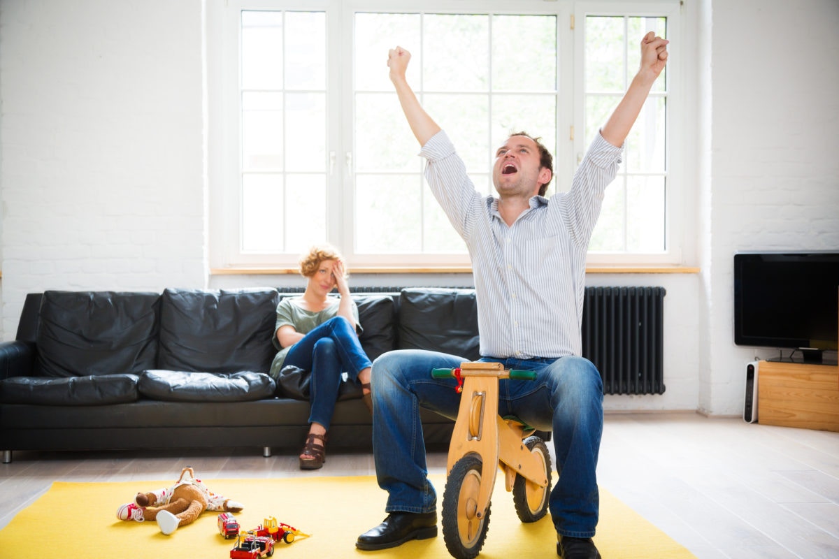 Husband is playing with kids toy bicycle while wife is looking strange at him