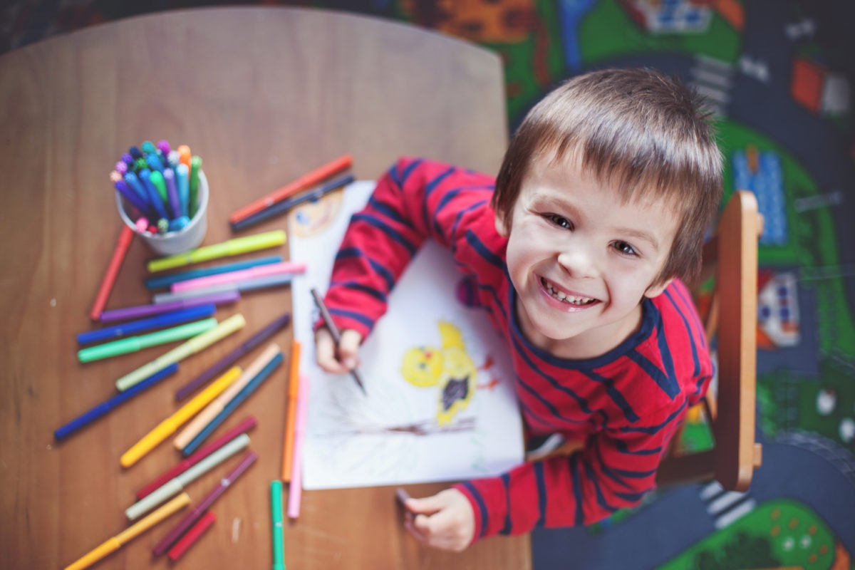little boy looking at camera, smiling while painting picture sitting at table in art class