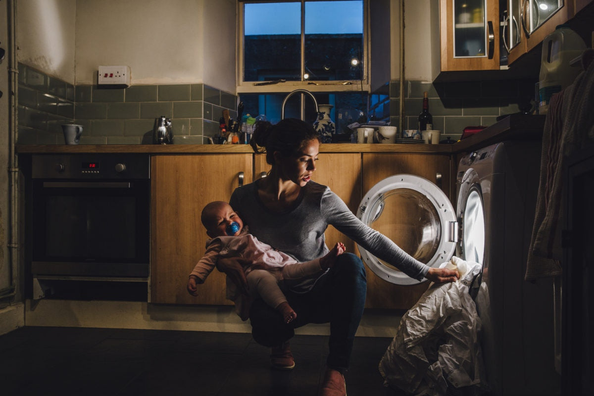 young mother is holding a newborn boy in her arms while putting laundry into the washing machine