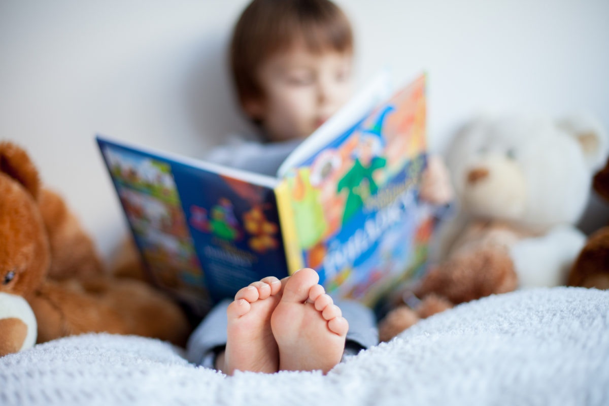 boy sitting on a bed reading story book surrounded with toys