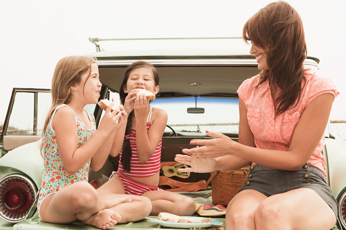mother with two girls sitting in car trunk and having breakfast