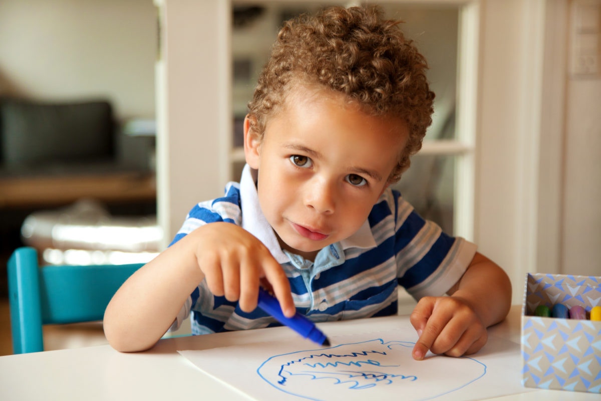 young boy drawing with crayons on sheets of white paper on a table