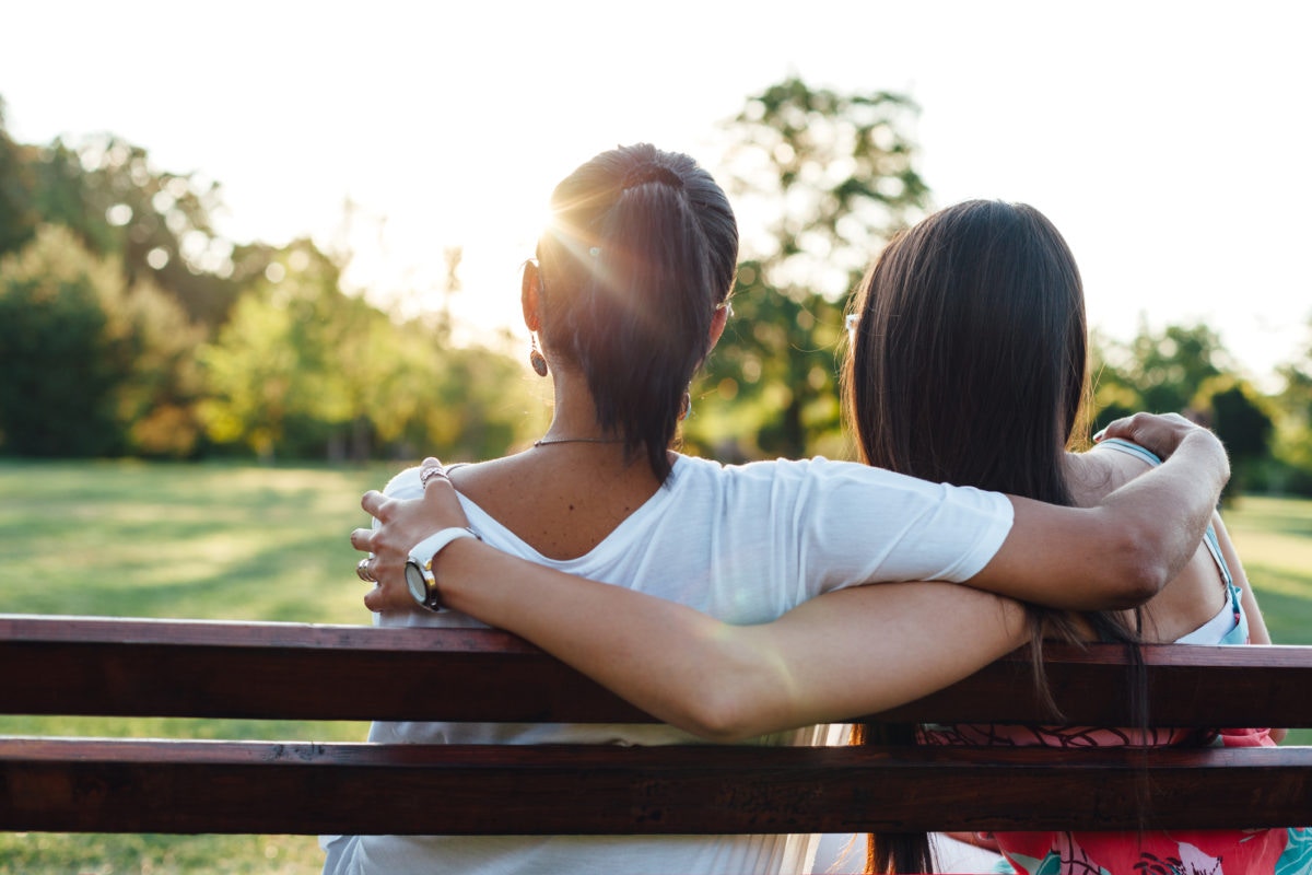 Back view of two women's hugging while sitting on a bench at the park