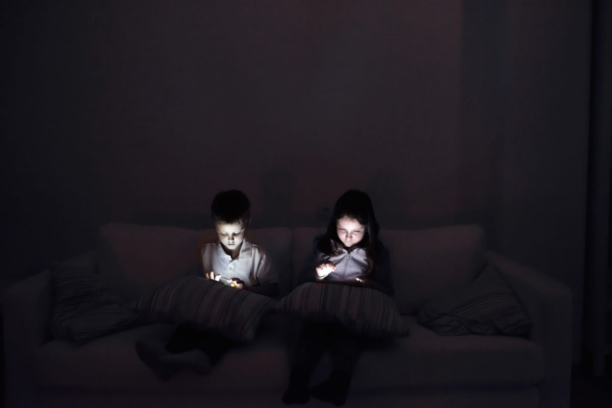 Little boy and girl playing with smart phone, sitting on sofa in dark room at night.