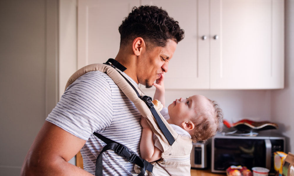 a father with small toddler son in carrier in kitchen indoors at home
